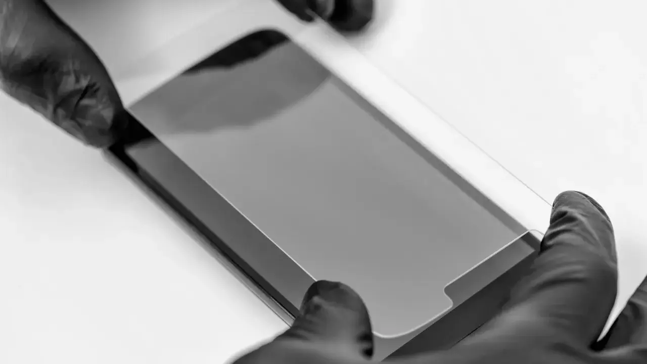 Ręce w czarnych rękawiczkach precyzyjnie nakładają szkło prywatyzujące na ekran smartfona, chroniąc prywatność.