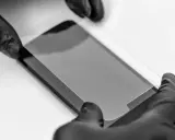 Ręce w czarnych rękawiczkach precyzyjnie nakładają szkło prywatyzujące na ekran smartfona, chroniąc prywatność.