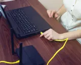 Ręka podłącza żółty kabel do laptopa, pokazując, czy można podłączyć monitor do laptopa. Obok leży router.