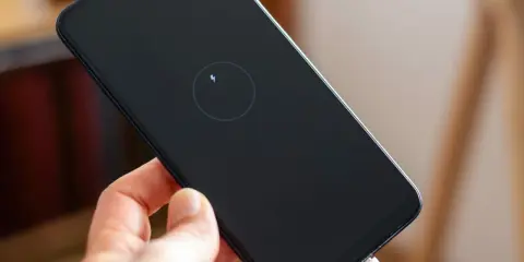Dłoń trzyma smartfon z ikoną ładowania zwrotnego na czarnym ekranie. Podłączony biały kabel USB.