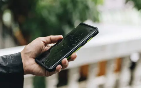 Dłoń trzyma mokry smartfon ip67 na tle zieleni. Krople deszczu spływają po ekranie.