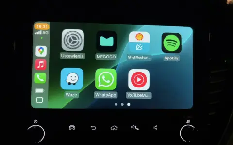Ekran samochodu z ikonami aplikacji: Ustawienia, MEGOGO, Shell Recharge, Spotify, Waze, WhatsApp, YouTube Music. Działa system iOS CarPlay.