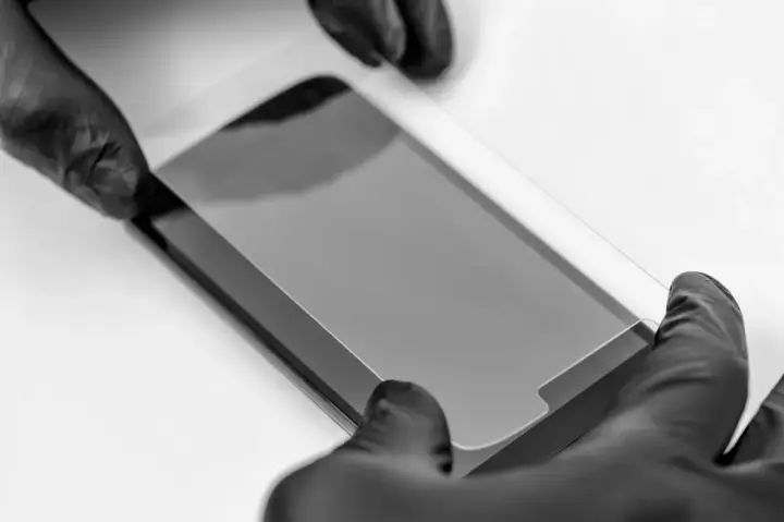 Ręce w czarnych rękawiczkach precyzyjnie nakładają szkło prywatyzujące na ekran smartfona, chroniąc prywatność.